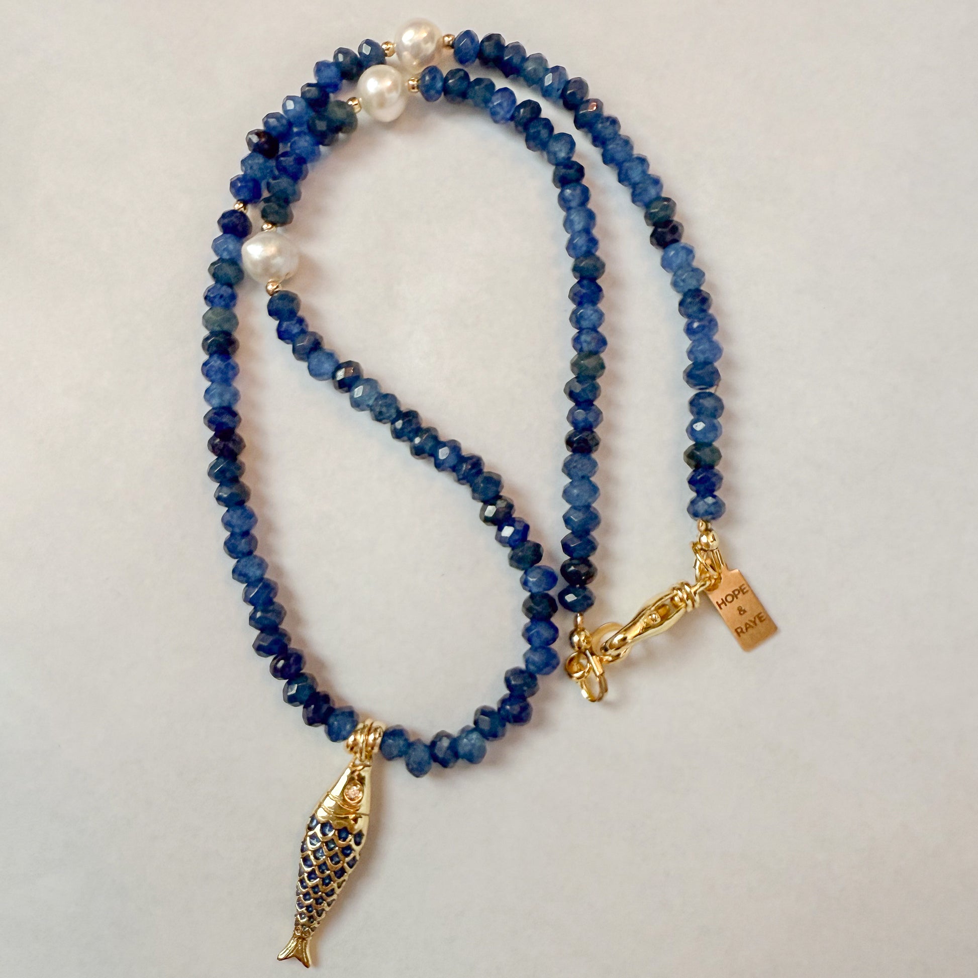 Tiny blue beaded natural gemstone necklace with gold-filled fish charm and gold-filled clasp. The piece features accents of natural freshwater pearls and is handcrafted by Hope and Raye in Atlanta, Georgia.